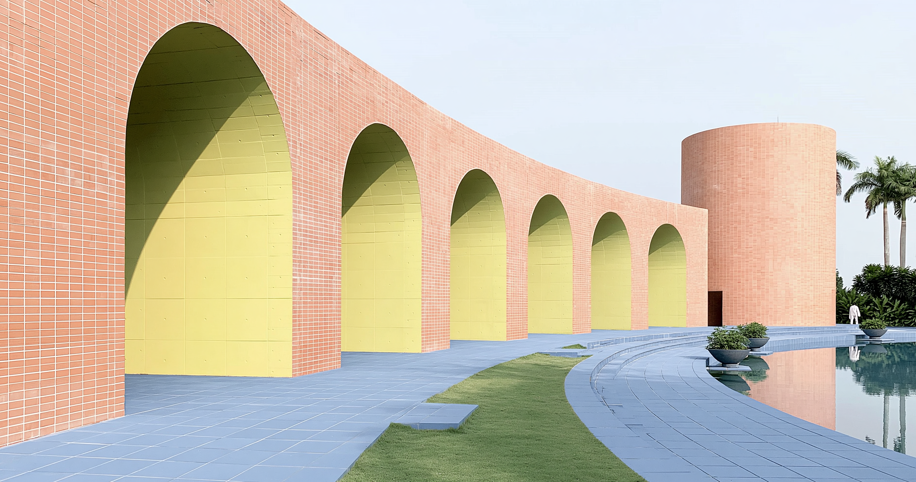 A long coral arcade curves beside a reflecting pool toward a cylindrical tower