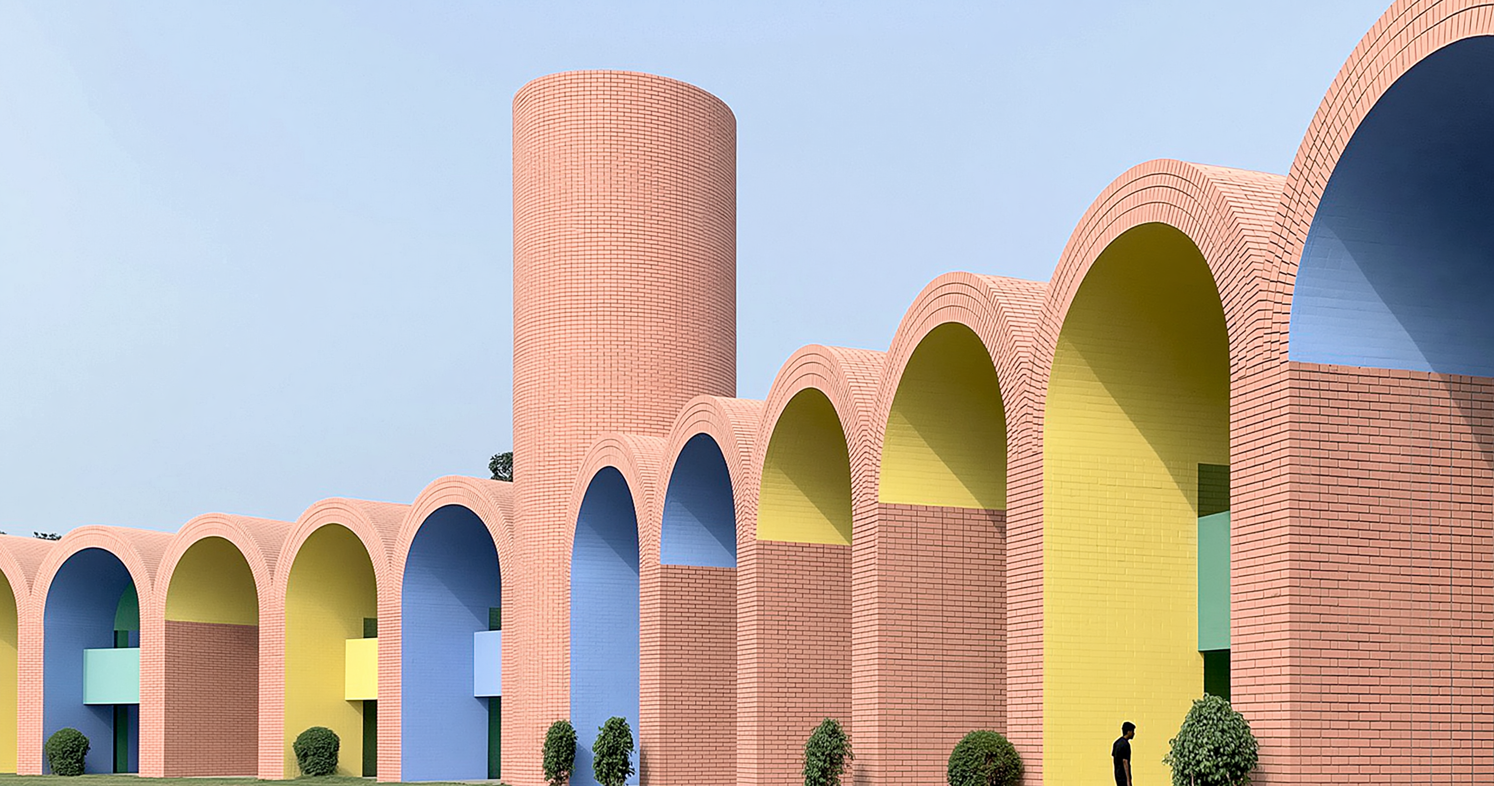 A tall cylindrical brick tower and a row of brick arches with yellow and blue interiors against a pale blue sky